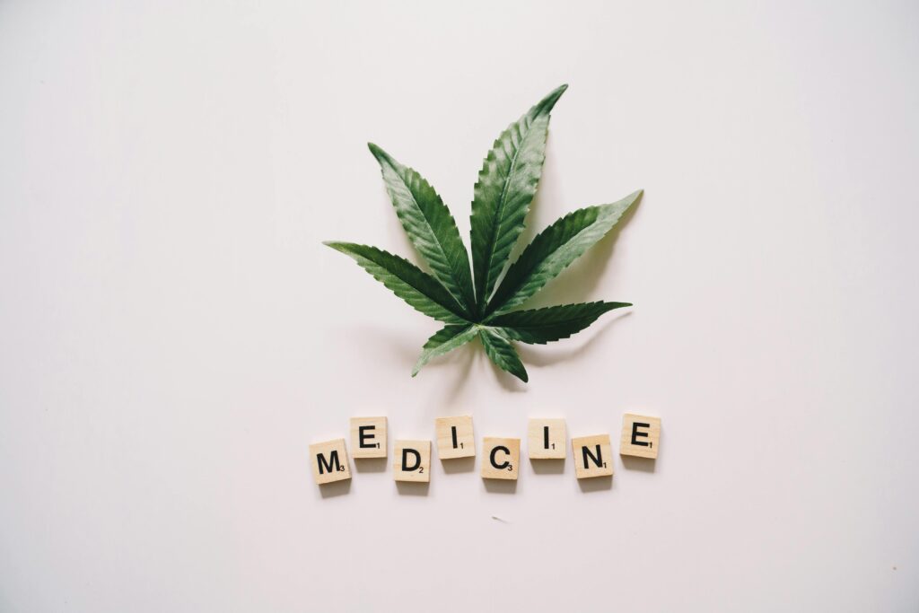 Flat lay of cannabis leaf and 'MEDICINE' spelled with Scrabble tiles on white background.